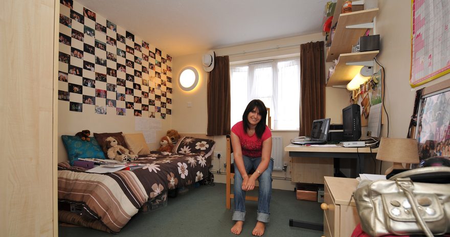 A standard ensuite featuring a student sitting on a chair, a single bed, a study desk and shelves and
