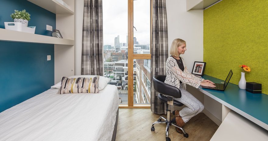 The classic ensuite bedroom with a bed, shelves, and a student at the desk using a computer at Urbanest Hoxton in London
